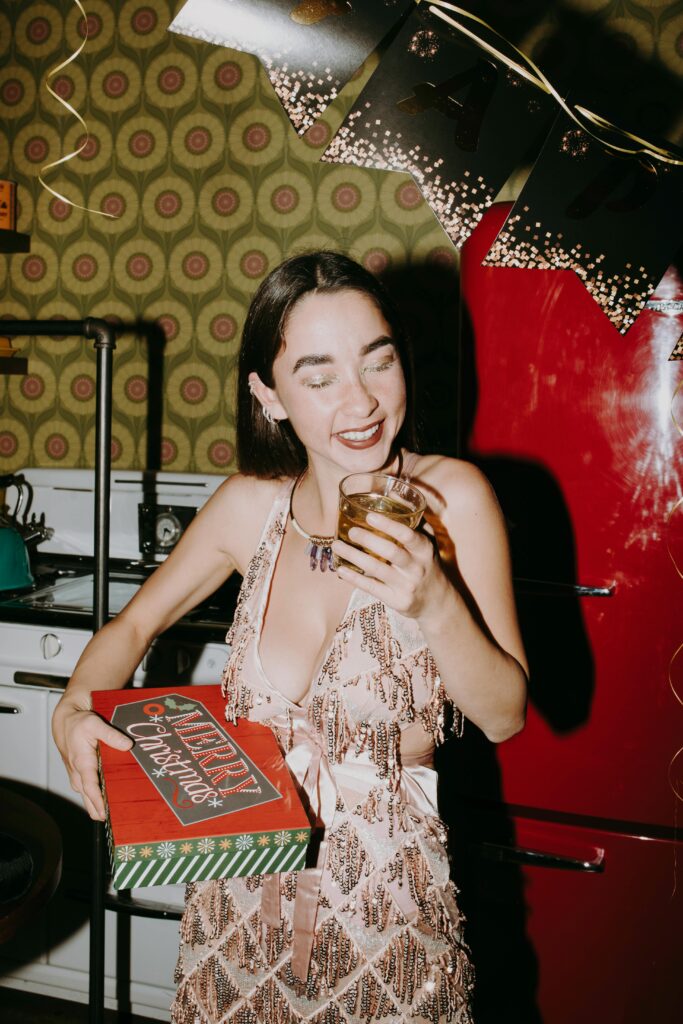 Smiling woman in festive dress with drink, celebrating in a retro kitchen.
